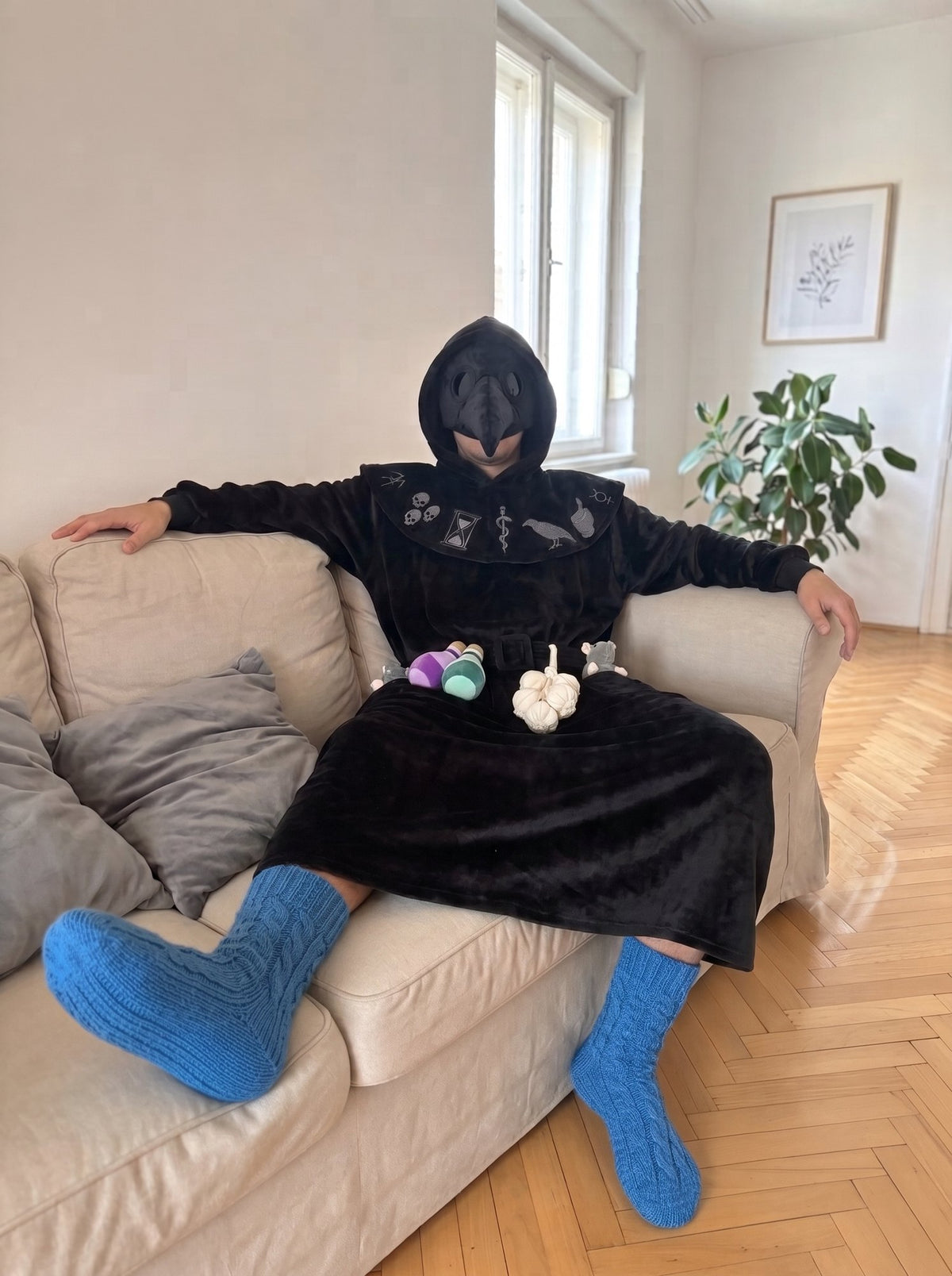 Man relaxing on a couch in the black Plague Doctor hooded blanket robe with beak mask, potions and garlic